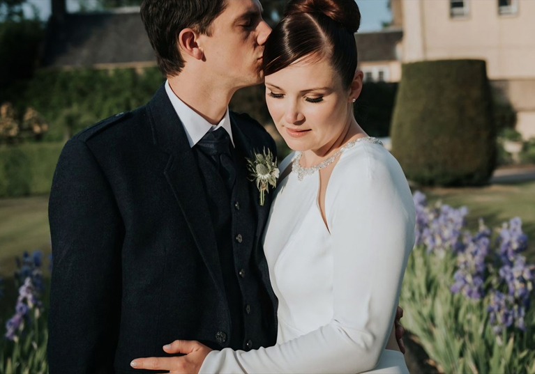 Groom kisses brides head as she looks down in gardens of their wedding venue, brides makeup by Jennifer Louise Studio.